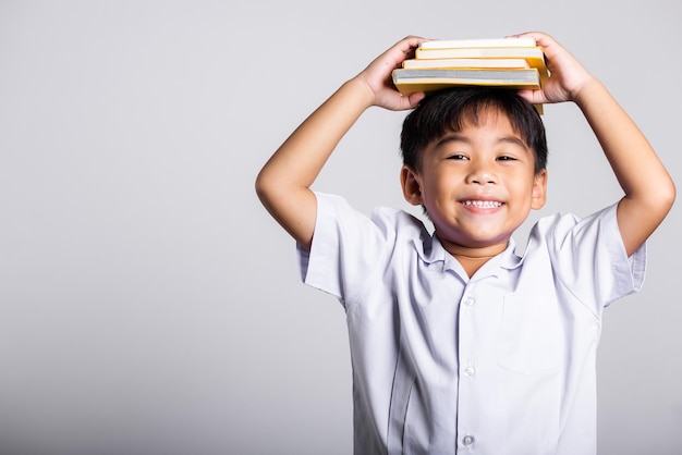 Asiatisches entzückendes kleinkind, das glücklich lächelt, trägt studententhaiuniform rote hosen, die ein buch über dem kopf halten und schreien