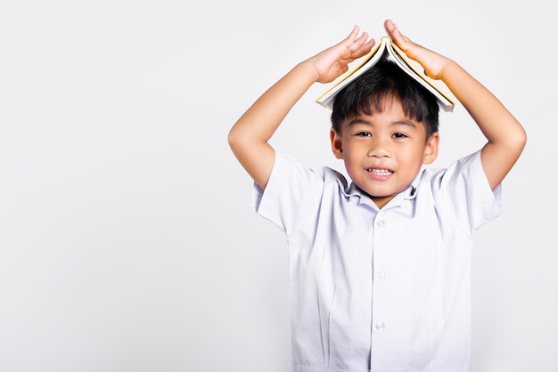 Asiatisches entzückendes Kleinkind lächelt glücklich Student Thai Uniform rote Hose stehen Buch über dem Kopf wie Dach im Studio geschossen isoliert auf weißem Hintergrund Porträt kleine Kinder Junge Vorschule