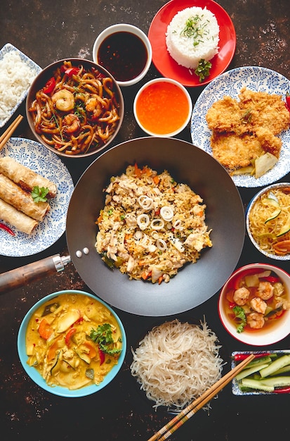 Asiatisches Essen Set Nudeln gebratener Reis mit Hühner Tom Yum Suppe Frühlingsrollen gebratener Fisch und Udon mit Garnelen Top-View auf dunklem rustikalen Hintergrund Oriental-Stil Essen Konzept Zusammensetzung