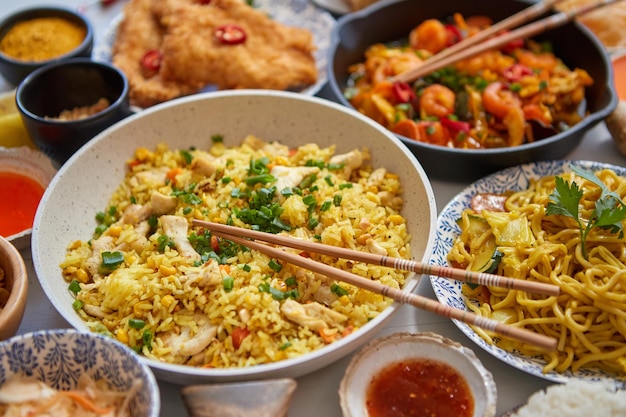 Asiatisches Essen wird serviert. Teller, Pfannen und Schüsseln voller leckerer orientalischer Gerichte, Nudeln, Hähnchenpfanne und Gemüsezutaten mit Gewürzsaucen und Essstäbchen auf weißem Holzhintergrund