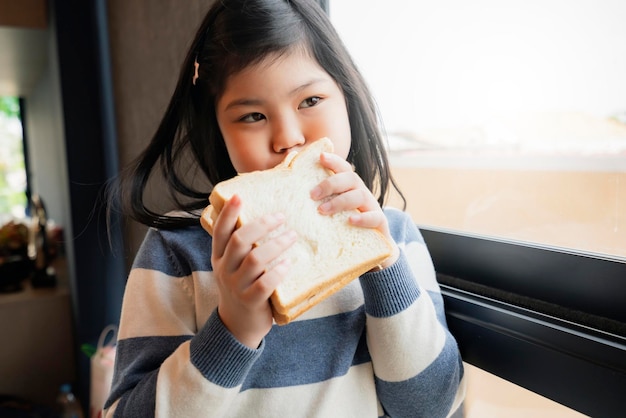 Asiatisches gesundes Mädchen im blauen und weißen Pullover genießen und fühlen sich gut mit Brotsandwisch in der Küche