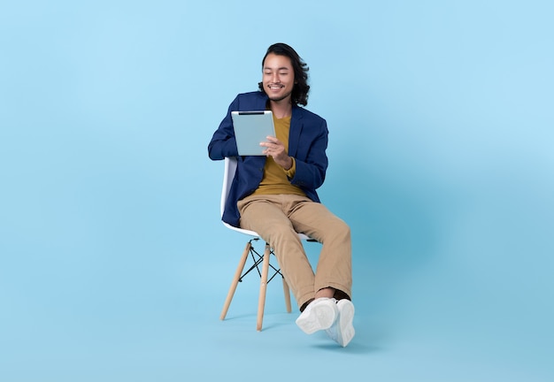 Asiatisches glückliches Lächeln des Geschäftsmannes mit einem digitalen Tablet beim Sitzen auf dem Stuhl lokalisiert auf hellem blauem Hintergrund.