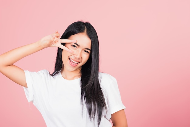 Asiatisches, glückliches Porträt, schöne, süße junge Frau, Teenager-Lächeln, das Finger zeigt, die V-Zeichen-Siegessymbol in der Nähe des Auges machen, das zur Kamerastudioaufnahme lokalisiert auf rosa Hintergrund mit Kopienraum schaut