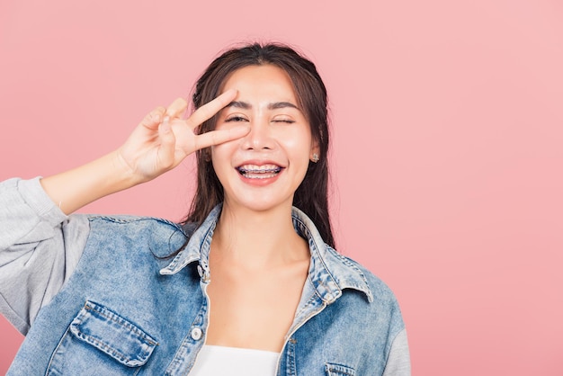 Asiatisches glückliches Porträt schöne süße junge Frau trägt Denim-Lächeln, das Finger zeigt, die V-Zeichen-Siegessymbol in der Nähe des Auges machen, das zur Kamerastudioaufnahme lokalisiert auf rosa Hintergrund mit Kopienraum schaut