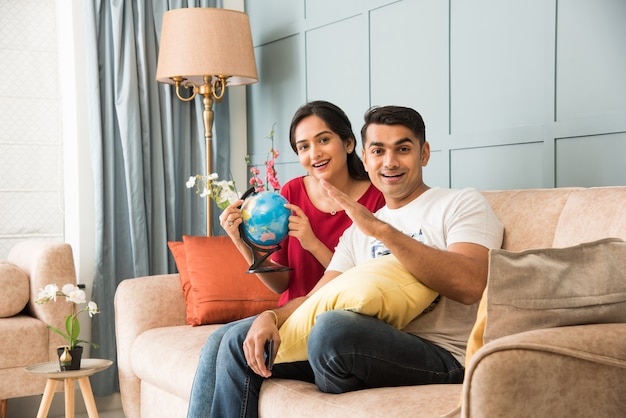 Asiatisches indisches junges Paar, das eine Urlaubsreise mit 3D-Globe-Modell plant, während es auf dem Sofa oder der Couch sitzt