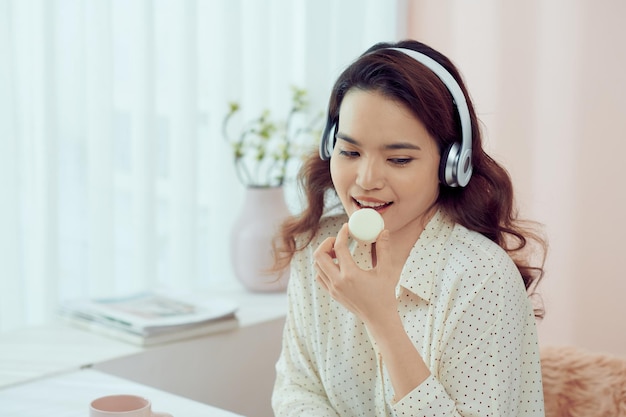 Asiatisches junges Mädchen mit Kopfhörern, das Macarons isst und Bücher im Wohnzimmer liest