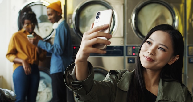 Asiatisches junges schönes fröhliches Mädchen lächelnd und posierend zur Smartphone-Kamera beim Selfie-Foto im Wäscheservice. Ziemlich glückliche Frau, die Selfies Fotos mit Telefon an Waschmaschinen macht.