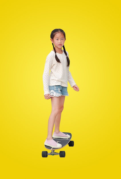 Asiatisches Kind des kleinen Mädchens, das auf einem Skateboard lokalisiert auf gelbem Hintergrund skatet. Kinderreiten auf Skateboard. Bild mit Beschneidungspfad