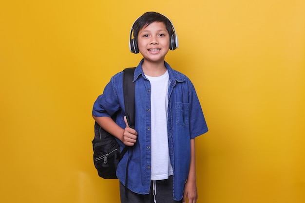 Asiatisches Kind im lässigen Denim-Stil mit Rucksack und kabellosen Kopfhörern, die in die Kamera lächeln