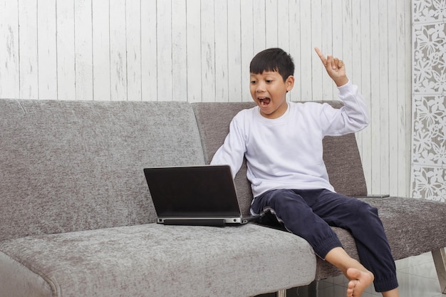 Asiatisches Kind im weißen Pullover ist aufgeregt und hebt die Hand, während es zu Hause mit dem Laptop online lernt