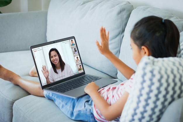 Asiatisches Kindermädchen, das auf dem Sofa sitzt und Videokonferenz-Chat mit Lehrer und Klassengruppe hat. Das Kind lernt während der Quarantäne aufgrund des Ausbruchs des Coronavirus COVID-19 von zu Hause aus.