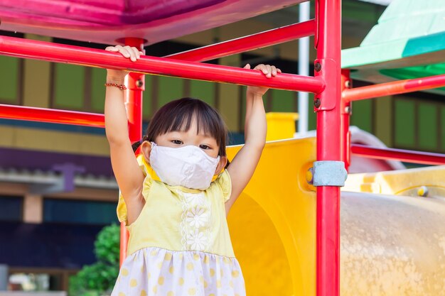 Asiatisches Kindermädchen, das eine Stoffgesichtsmaske trägt, wenn sie ein Spielzeug am Spielplatz spielt. Soziale Distanz.