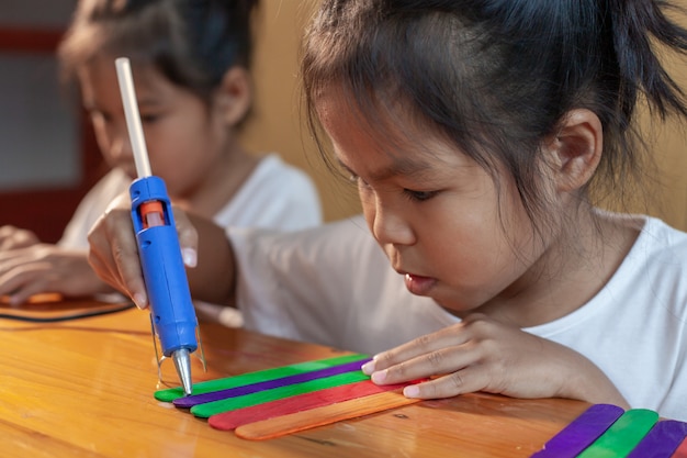 Asiatisches Kindermädchen, das farbige Eiscremesticks durch heiße elektrische Schmelzklebepistole klebt. Kinder haben Spaß daran, ein Handwerksprojekt zu bauen.