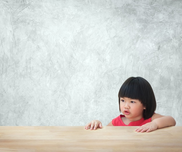 Asiatisches Kindermädchen, das hinter leerer Tabelle mit Betonmauer sitzt