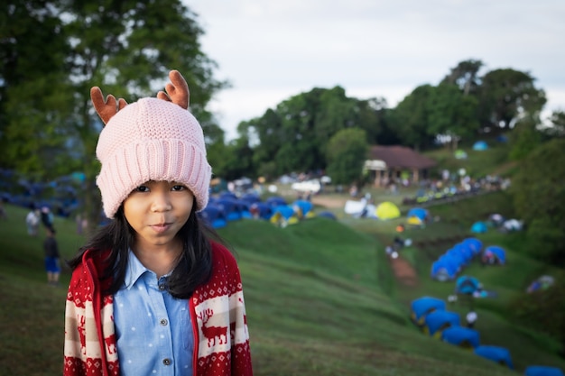 Asiatisches Kindermädchen, das warmen Hut trägt, der auf dem Hügel mit grünem Gras steht
