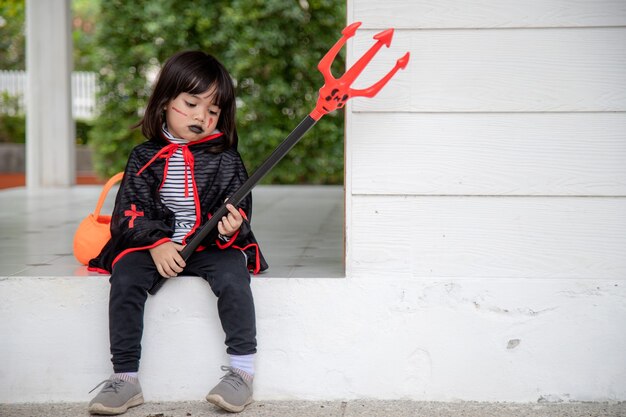 Asiatisches Kindermädchen im Dämonenkostüm, das schwarzen und roten Dreizack hält, glückliches Halloween-Konzept