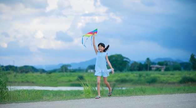 Asiatisches Kindermädchen mit einem Drachen, der auf Wiese im Sommer in der Natur läuft und glücklich ist