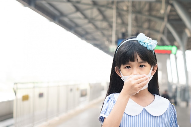 Asiatisches kleines junges Mädchen, das Maske trägt, um Covid-19- oder Coronavirus-Infektion am Bahnhof zu verhindern.