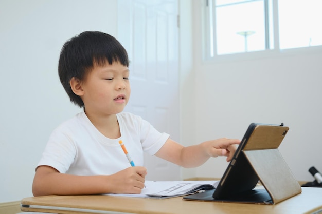 Asiatisches kleines Kind, das Tablet-Computer verwendet, um Informationen über das Internet zu suchen, das während des Online-Unterrichts zu Hause Hausaufgaben lernt