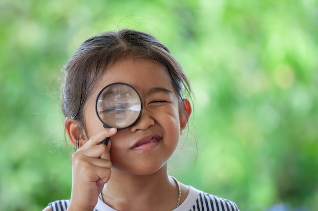 Asiatisches kleines Kindermädchen, das durch ein Vergrößerungsglas auf grünem Naturhintergrund schaut