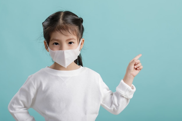 Asiatisches kleines Mädchen, das Atemschutzmaske trägt, um Coronavirus-Ausbruch zu schützen und Hand auf leeren Copyspace zu zeigen