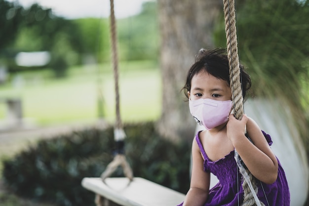 Asiatisches kleines Mädchen, das eine schützende Gesichtsmaske trägt, die auf einer Holzschaukel sitzt