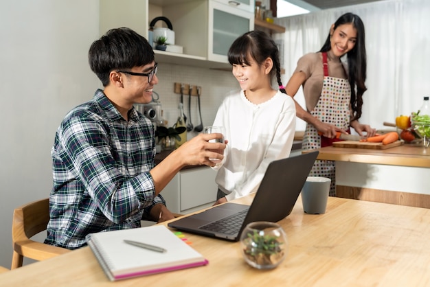 Asiatisches kleines Mädchen, das Papa hilft, mit Computer-Laptop zu arbeiten, während Mutter kocht