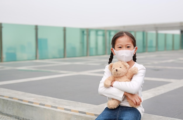 Asiatisches kleines Mädchen mit Maske zum Schutz vor Corona-Virus und Luftverschmutzung pm25 Staub mit umarmender Teddybärpuppe, die im Freien sitzt Coronavirus covid19 und Verschmutzungsschutzkonzept