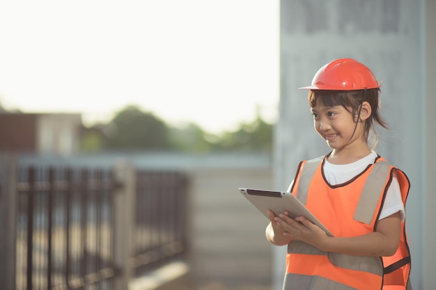 Asiatisches kleines Mädchen mit Schutzhelm auf dem Kopf und Tablet in der Hand auf einer Baustelle