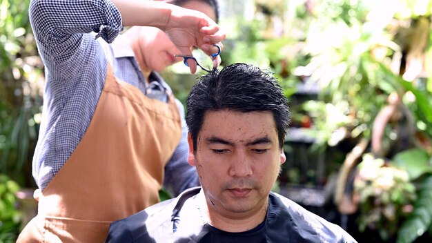 Asiatisches Liebespaar, das Haarschnitt macht, um sein Aussehen zu Hause aufzufrischen. Salon und Friseursalon im Garten.