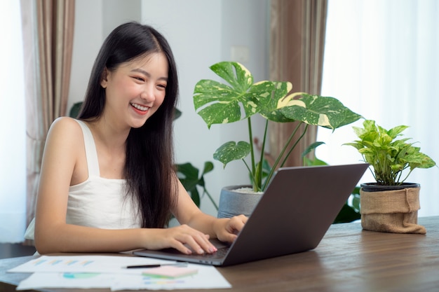 Asiatisches Mädchen, das Daten in ihr Computer-Notizbuch schreibt, um ihren Bericht über den Holzdeak mit der grünen Pflanze auf dem Tisch vorzubereiten