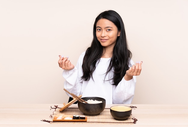 Asiatisches Mädchen des Teenagers, das asiatisches Essen auf beiger Wand isst, die Geldgeste macht