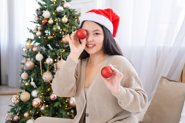 asiatisches mädchen in weihnachtsmütze, das spaß hat und rote weihnachtskugeln hält, die zu hause auf dem bett sitzen, weihnachten ti