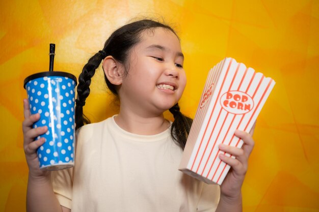 Asiatisches Mädchen isst Popcorn und trinkt Limonade auf gelbem Wandhintergrund Kinokonzept