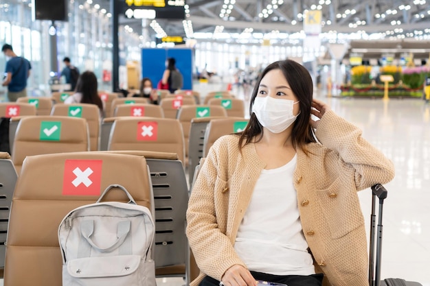 Asiatisches Mädchen mit Maske sitzt am internationalen Flughafen, während es auf den offenen Check-in-Schalter wartet Soziale Distanz an öffentlichen Orten Sitzordnung im Flughafenbereich Neues normales Lifestyle-Konzept