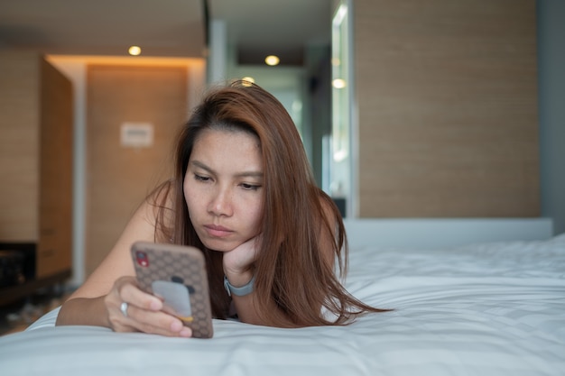 Asiatisches Mädchen mit Smartphone, das auf dem Bett liegt