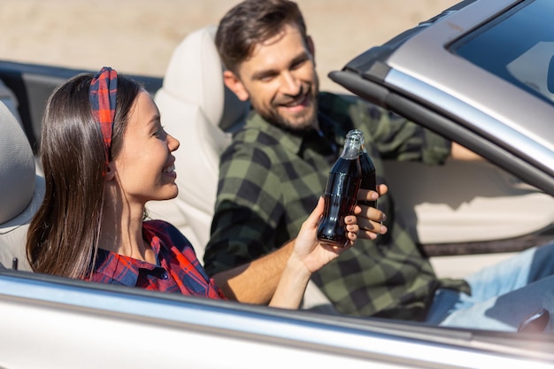 Asiatisches Mädchen und Kerl, die mit Koksflaschen klicken, die im Cabrio sitzen