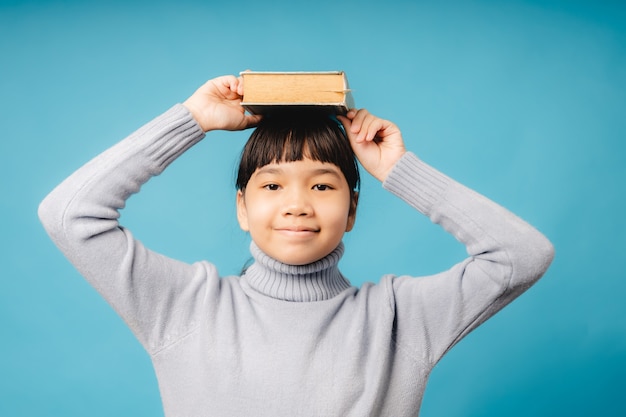 Asiatisches Mädchenkind, das ein Lehrbuch auf dem Kopf der kreativen Idee und des hellen Lernens hält, stellen sich vor, lernen und Bildungskonzept