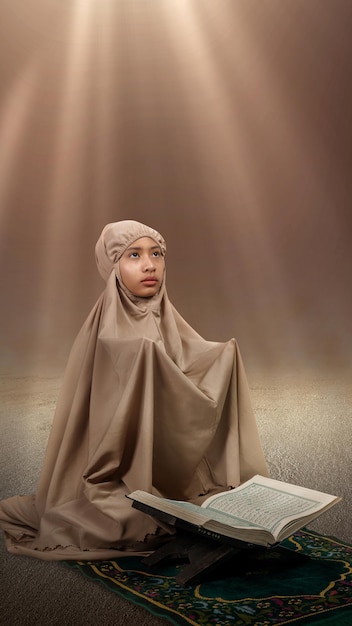 Asiatisches muslimisches Mädchen in einem Hijab mit Koran auf dem Gebetsteppich hob die Hände und betete