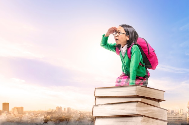 Asiatisches nettes Mädchen mit den Gläsern und Rucksack, die auf dem Stapel von Büchern mit Stadt und blauem Himmel sitzen