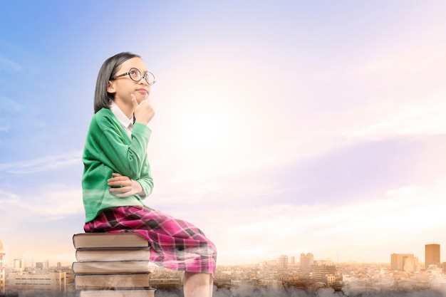Asiatisches nettes Mädchen mit Gläsern denken beim Sitzen auf dem Stapel von Büchern mit Stadt und blauem Himmel