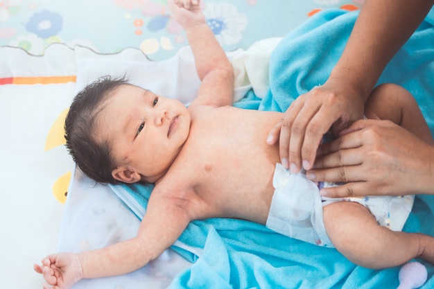 asiatisches neugeborenes Baby, das sich auf ihrem Bett hinlegt und ihre Mutter schaut, während sie ihre Windel ändert