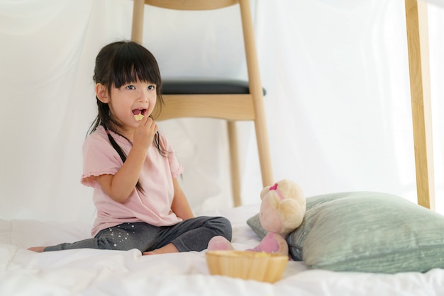 Asiatisches niedliches kleines Mädchen, das Snack isst, während in einer Deckenfestung im Wohnzimmer zu Hause sitzen