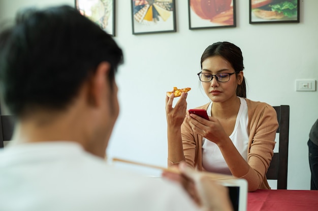 Asiatisches Paar, das zusammen Instant-Nudeln und Pizza in der Küche isst. Genießen Sie eine gesunde Mahlzeit. Lebensstil zum Abendessen und bleiben Sie zu Hause. Frau, die während des Essens smarphone für Suchinformationen verwendet.