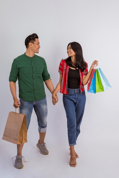Asiatisches Paar hält Händchen und schaut einander an, während es die Einkaufstüten auf isoliertem Hintergrund mitbringt