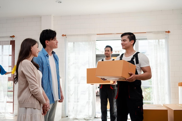 Asiatisches Paar prüft beim Entladen von Kisten und Möbeln aus einem Pickup in ein neues Haus mit Dienstfracht zwei Männer Umzugsarbeiter in Uniform hebende Kisten Konzept des Umzugs und der Lieferung zu Hause