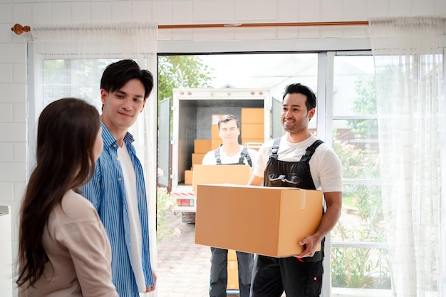 Asiatisches Paar prüft beim Entladen von Kisten und Möbeln aus einem Pickup in ein neues Haus mit Dienstfracht zwei Männer Umzugsarbeiter in Uniform hebende Kisten Konzept des Umzugs und der Lieferung zu Hause