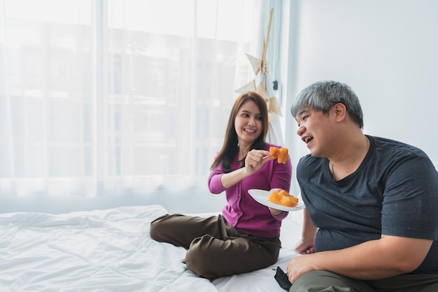 Asiatisches Paar sitzt auf einem weißen Bett im Haushalt und isst Donuts als Snack