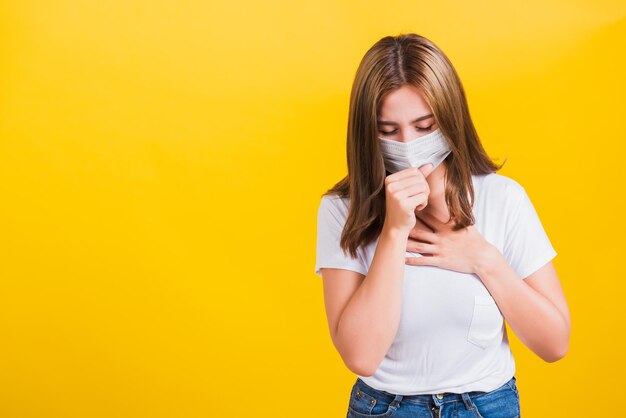 Asiatisches Porträt schöne süße junge Frau stehend tragen T-Shirt Husten in Maske Schutz vor COVID-Virus-Epidemie oder Luftverschmutzung isoliert, Studioaufnahme auf gelbem Hintergrund mit Kopierraum