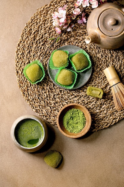 Asiatisches Reisdessert süßer grüner Matcha-Mochi mit Tasse geschäumtem Matcha-Tee und grünem Matcha-Pulver in Keramikplatte auf braunem Tisch Teekanne und frühlingsrosa blühender Zweig auf gestrickter Strohserviette